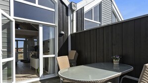 Een terras buiten met een ronde tafel en rieten stoelen, een schuifdeur naar een binnenruimte, en een donker houten hek.