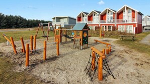 Een speeltuin met schommels en klimconstructies, een picknicktafel en een gebouw met rode en witte gevels op de achtergrond.