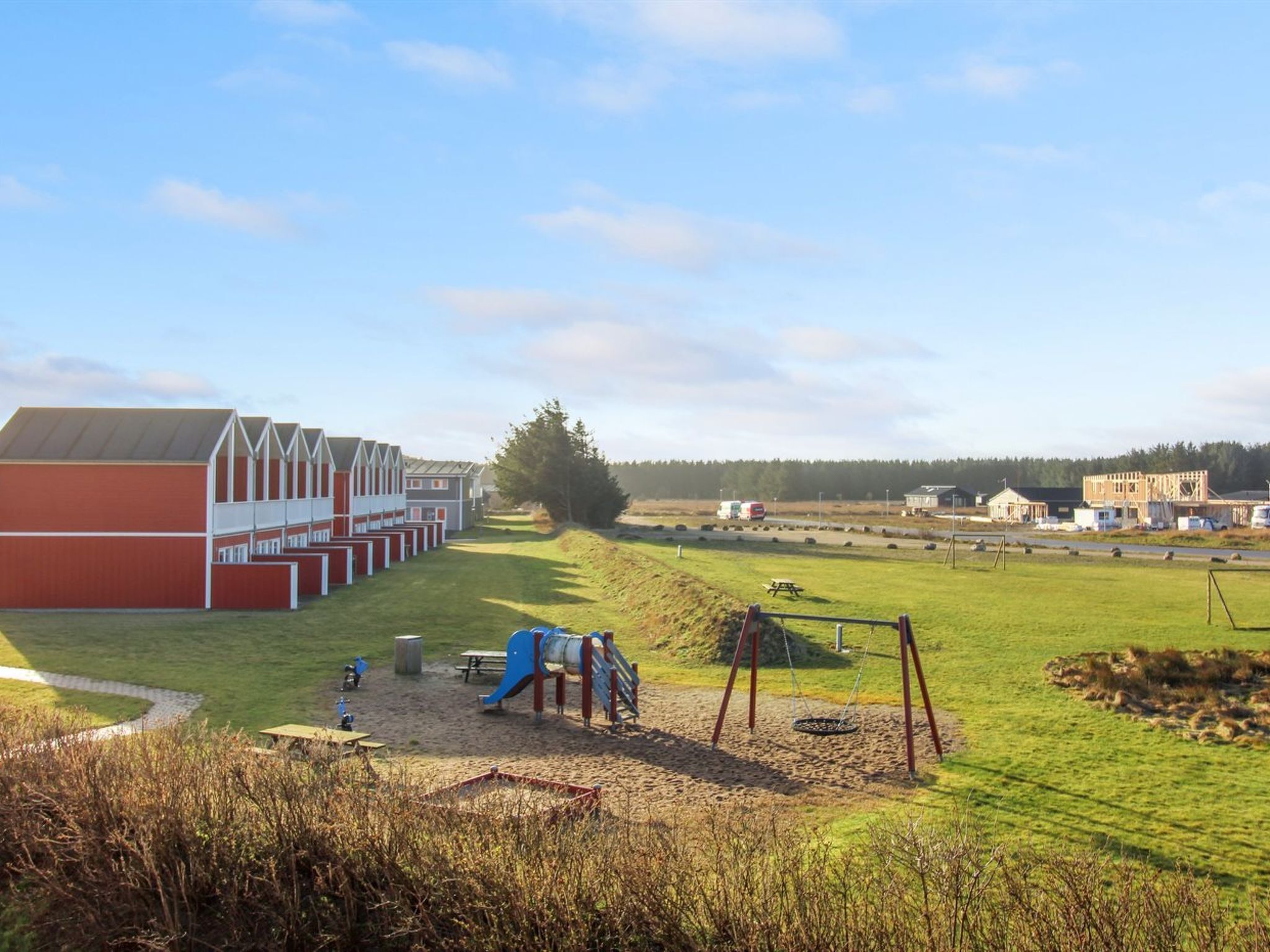 A playground with swings and a slide, a red building with white trim, and a grassy field with a few trees.