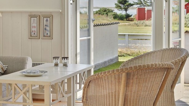 A room with a white table and wicker chair, framed pictures on the wall, and a view of a grassy area and a red building outside.