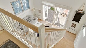 A spacious room with a high ceiling, a wooden staircase, a balcony with a view, and a cozy seating area with a sofa and cushions.