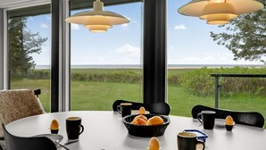Een eetgedeelte met een tafel gedekt voor twee personen, grote ramen met uitzicht op een grasveld en strand, en hanglampen erboven.