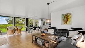 Een moderne woonkamer met een zwarte leren bank, een houten salontafel en een eethoek met rieten stoelen.