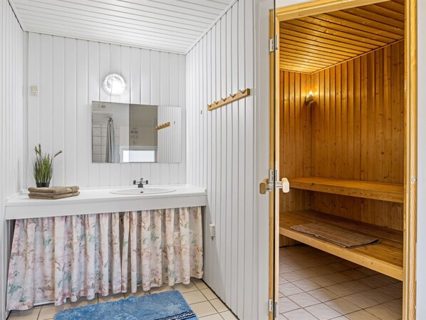 Un baño con tocador blanco, espejo y sauna de madera visible a través de una puerta abierta.