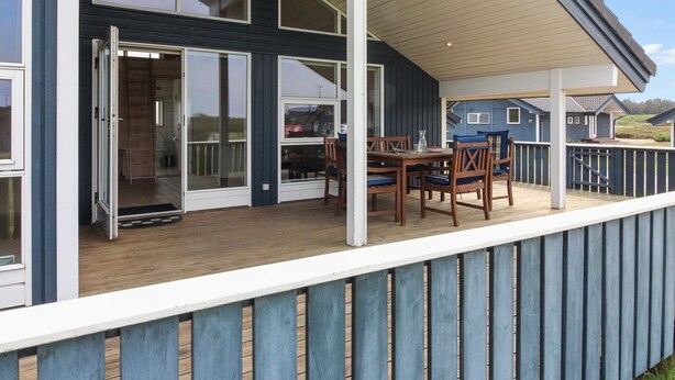 Una terrazza esterna coperta con tavolo e sedie in legno, una porta scorrevole in vetro che dà su un ambiente interno e una ringhiera blu.