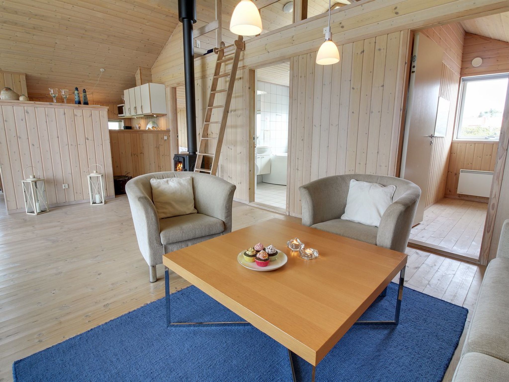 Un interior de cabaña acogedor con una mesa de comedor de madera, dos sillones, una zona de cocina con estufa y una escalera.