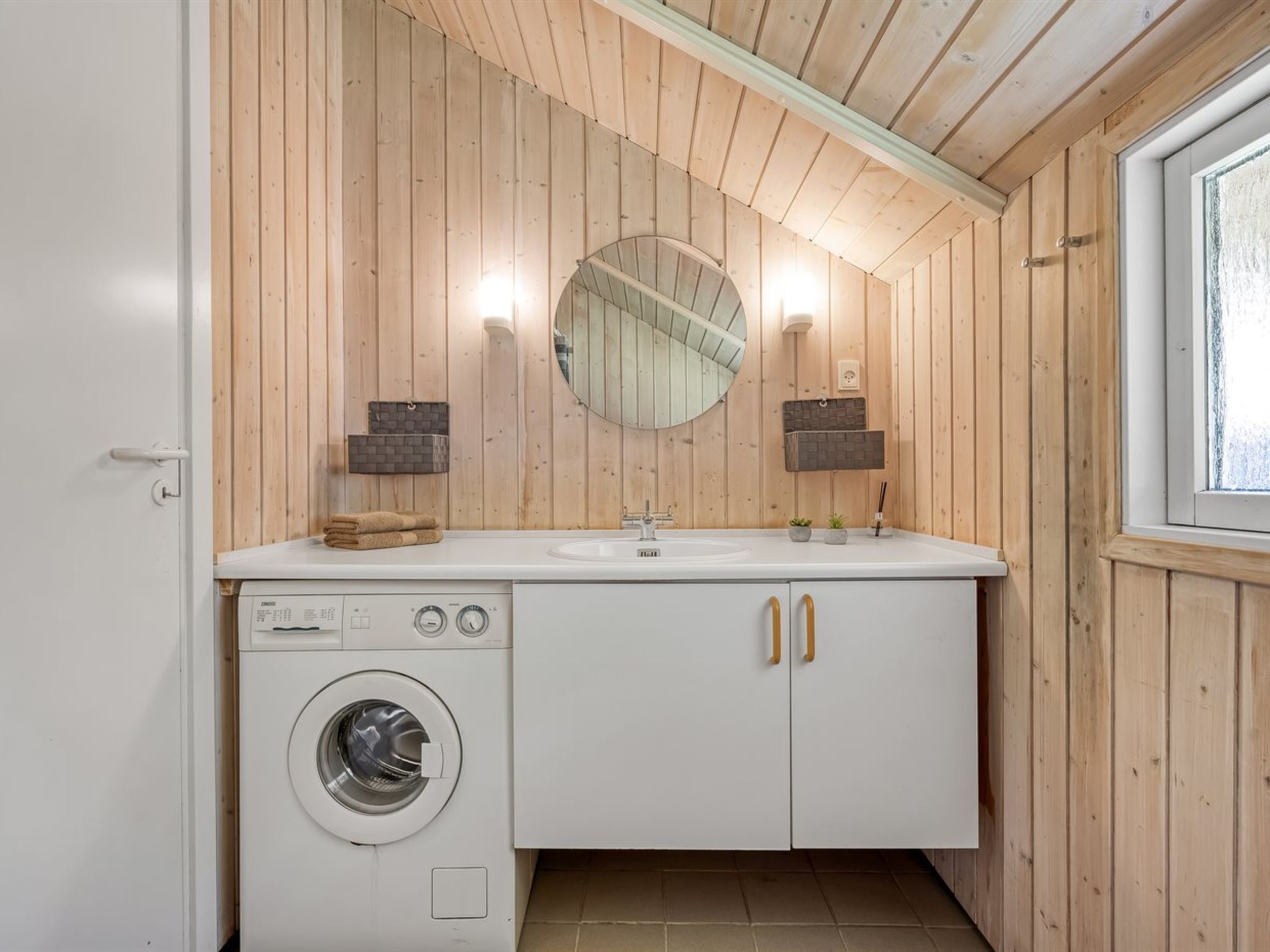 Een kleine badkamer met houten lambrisering, een ronde spiegel, een wastafel, een wasmachine en twee handdoekrekken.