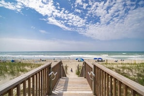 A wooden boardwalk leading to a beach with people and umbrellas. - Steps to Beach: Pools + Pickleball + Tennis (Emerald Isle)