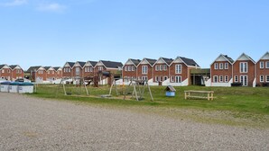 Eine Reihe von roten Backsteinhäusern mit weißem Zierdekor und Giebeldächern, ein Spielplatz mit Schaukeln und einer Rutsche sowie ein Kiesweg davor.