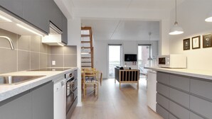 Eine moderne Küche mit grauen Schränken, einem eingebauten Backofen und einer Mikrowelle. Ein Holztisch mit Stühlen und eine Leiter zu einem Erkerraum sind ebenfalls vorhanden.