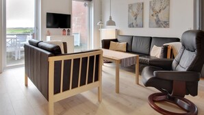 Ein modernes Wohnzimmer mit einem schwarzen Ledersofa, einem Holztisch und einem Leder-Sessel. Ein Fernseher steht auf einer weißen Kommode, und an der Wand hängen zwei Gemälde.