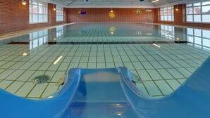 Una piscina interior con dos toboganes azules, suelo de baldosas y pared de ladrillo con ventanas.