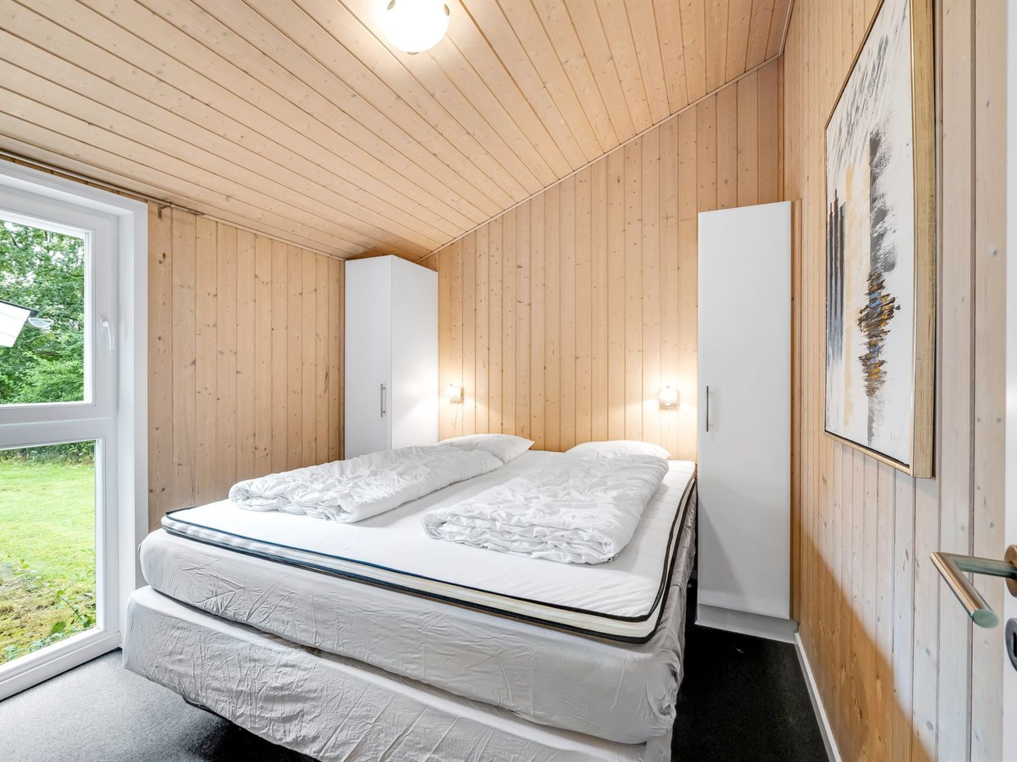A bedroom with a double bed, wooden walls, a wardrobe, and a painting on the wall.