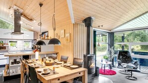 A modern kitchen with wooden cabinets, a dining area with a wooden table and chairs, a black stove, and a view of the outdoors through large windows.