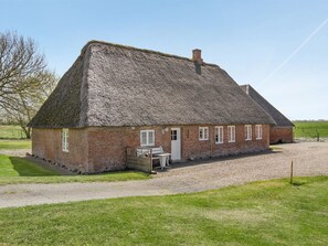 Ein Backsteinhaus mit einem Strohdach, einer Schotterauffahrt und einer kleinen Sitzgelegenheit im Freien.