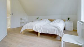 Quarto com teto inclinado, cama com roupa de cama branca, duas mesas de cabeceira com abajures e uma cadeira branca.