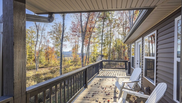 Um deck de madeira com cadeiras Adirondack, com vista para uma floresta com folhagem de outono e uma montanha ao longe.