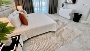 Un dormitorio moderno con una cama, lámparas de noche, un televisor, un tocador blanco y una planta en maceta.