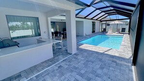 Un área de piscina exterior moderna con techo de vidrio, zona de estar con sillas y una pequeña barra.