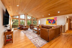 A spacious living room with wooden floors, large windows, a fireplace, and a mounted TV.