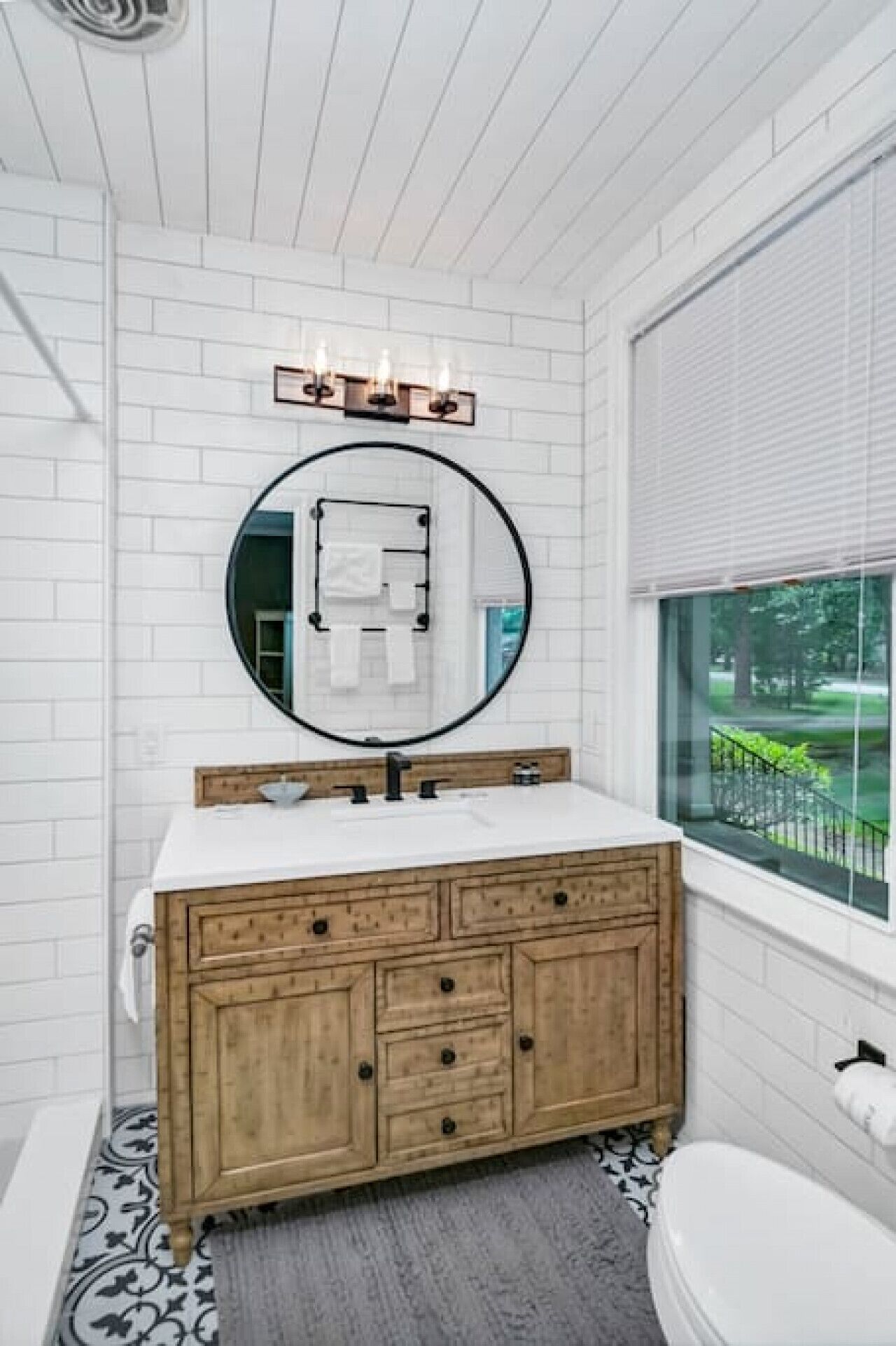 Un baño con azulejos blancos estilo metro, un tocador de madera con varios cajones, un espejo redondo y una ventana con vistas a zonas verdes.