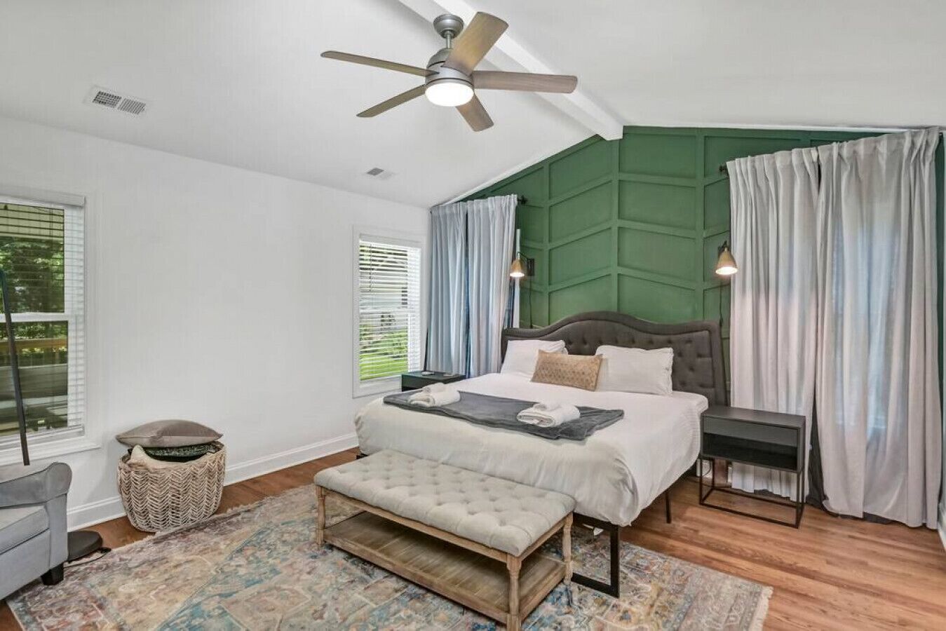A bedroom with a bed, bedside tables, a bench, a ceiling fan, and a window with curtains.