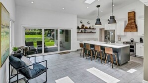 Une cuisine moderne dotée d’un grand îlot central, d’un éclairage suspendu et offrant une vue sur un jardin grâce à de grandes portes-fenêtres coulissantes.