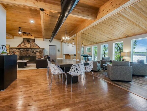 A spacious living area with a stone fireplace, wooden ceiling, and large windows offering a view of the outdoors. - Hygge Lodge is a gorgeous lakefront gathering place for year round family fun. (New Auburn)
