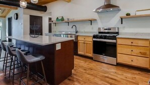 Una cocina moderna con una isla central, taburetes de bar, electrodomésticos de acero inoxidable, armarios de madera y una lámpara colgante.