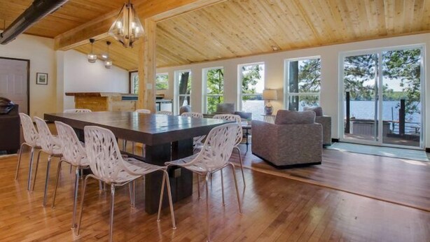 Una habitación amplia con una gran mesa de madera para comer rodeada de sillas, un sofá y vistas a un lago a través de amplios ventanales.