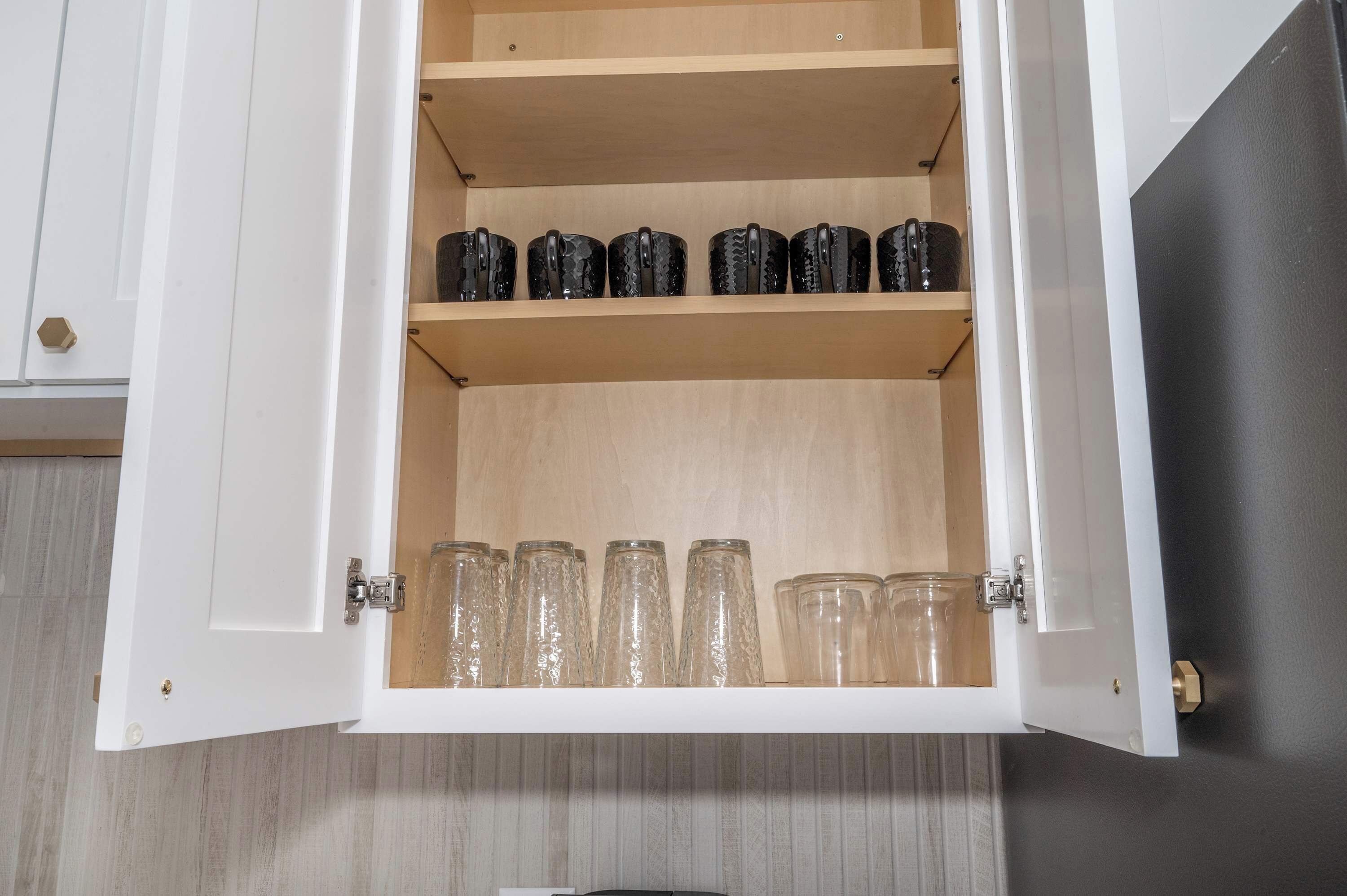 A kitchen cabinet with two shelves, one holding black mugs and the other clear glasses.