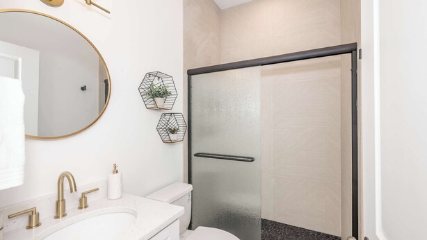 Ein modernes Badezimmer mit einem runden Spiegel, einem weißen Waschbecken mit goldfarbenen Armaturen, einer Toilette und einer Duschkabine aus Glas.