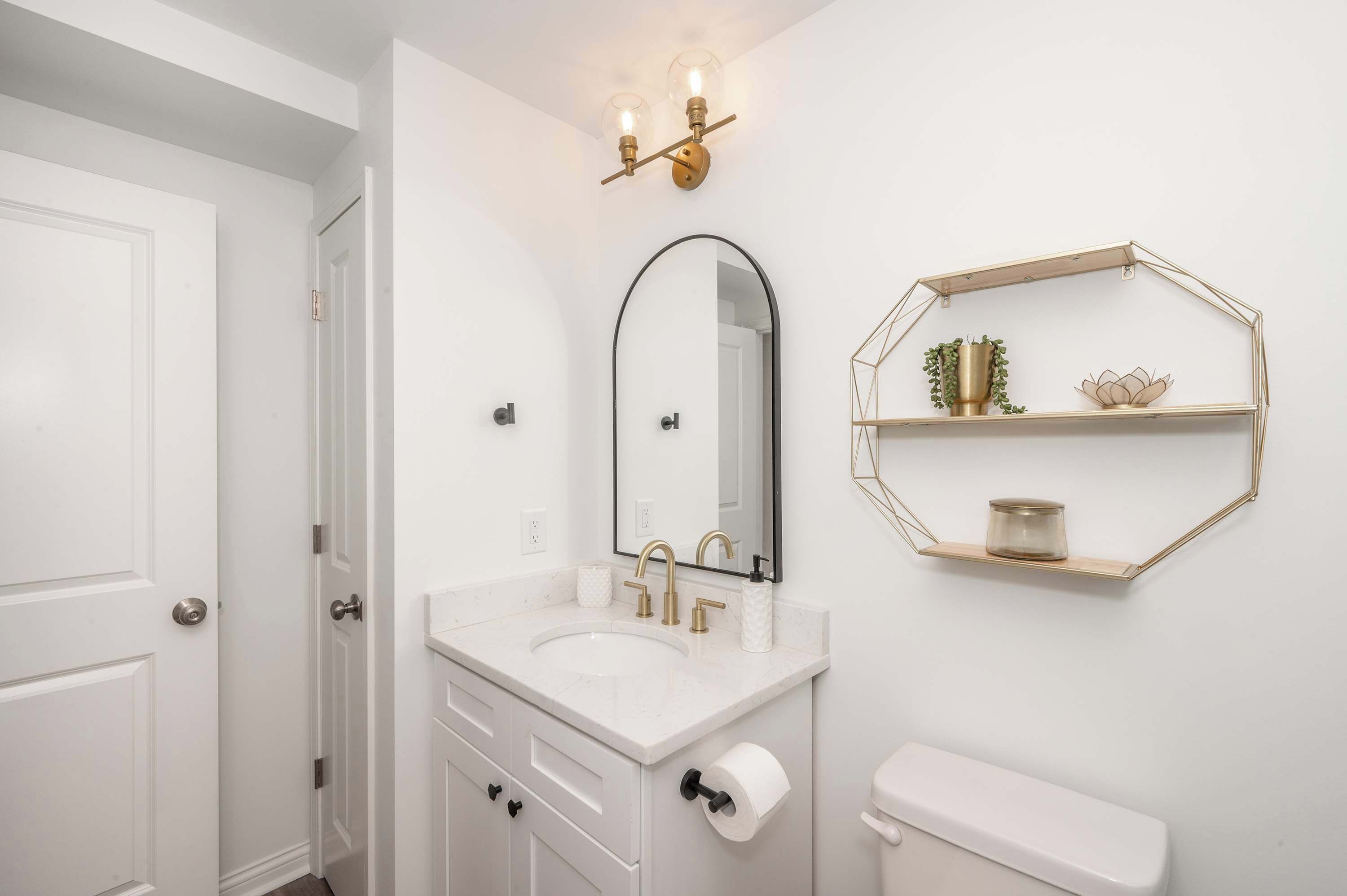 A bathroom with a white vanity, a sink, a toilet, a mirror, and a wall-mounted shelf with decorative items.