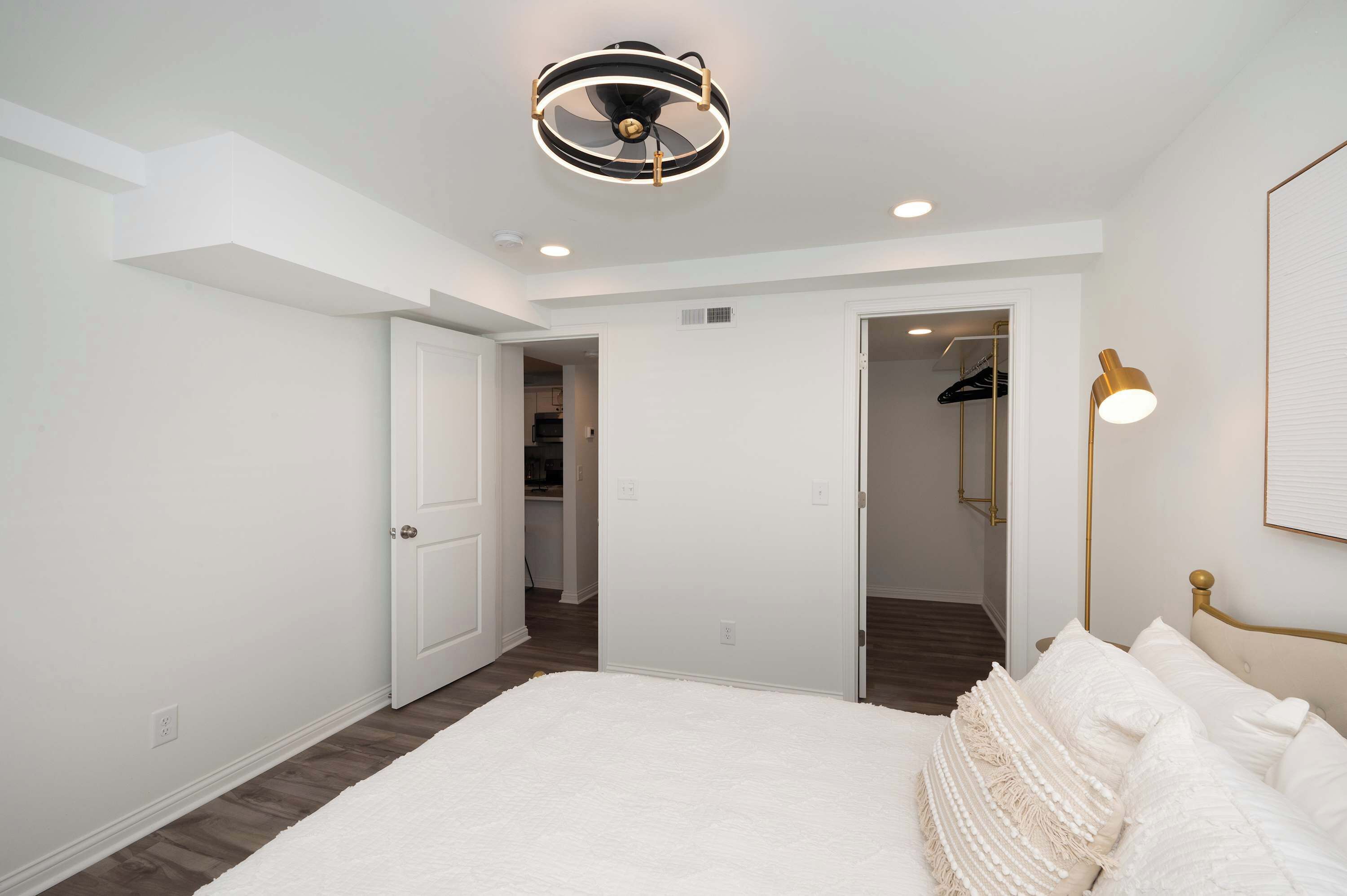A bedroom with a large bed, a ceiling fan, recessed lighting, and a hallway leading to a closet.