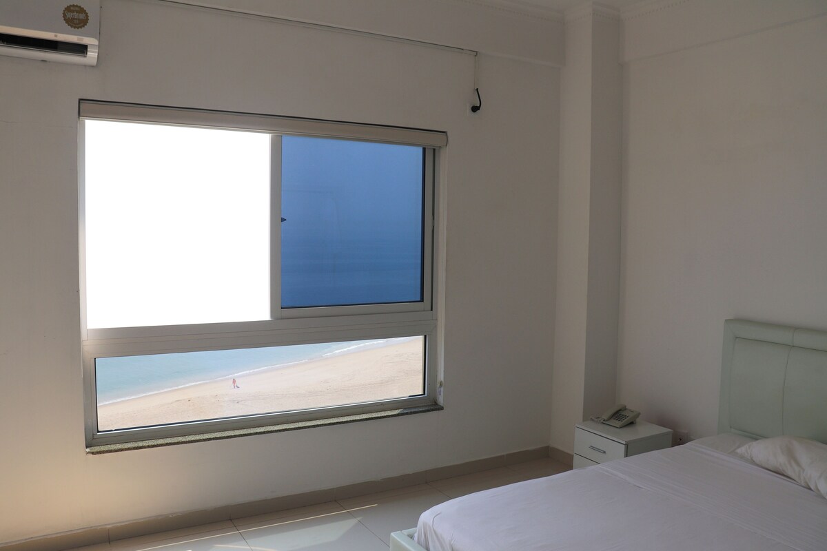 A bedroom with a bed, a nightstand, a phone, and a window with a beach view.