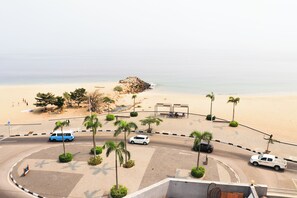 A beachside road with palm trees, a sandy beach, and a rocky outcrop. - Hotel Coral da Ilha (Luanda)