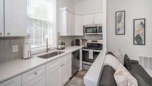 Una cocina moderna con armarios blancos, un microondas integrado y un horno. Hay una zona de comedor con un sofá gris y una mesita.