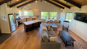 Una cocina amplia con una isla central, un comedor con una mesa redonda y sillas, una sala de estar con sofá y televisor, y un ventanal con vistas al exterior.