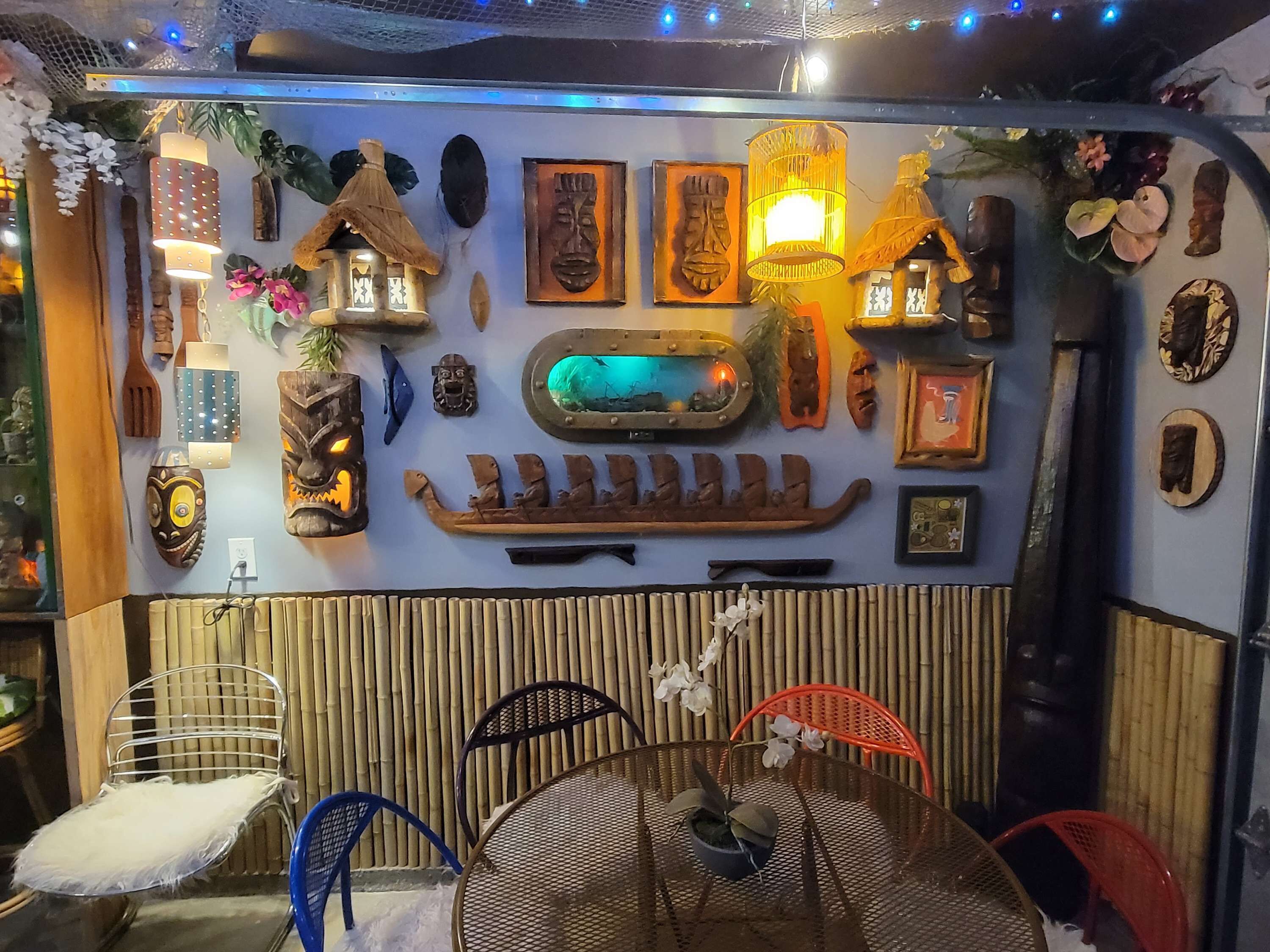 A room with a wall decorated with various cultural artifacts, including masks and a boat sculpture. There are several chairs and a table with a potted plant.