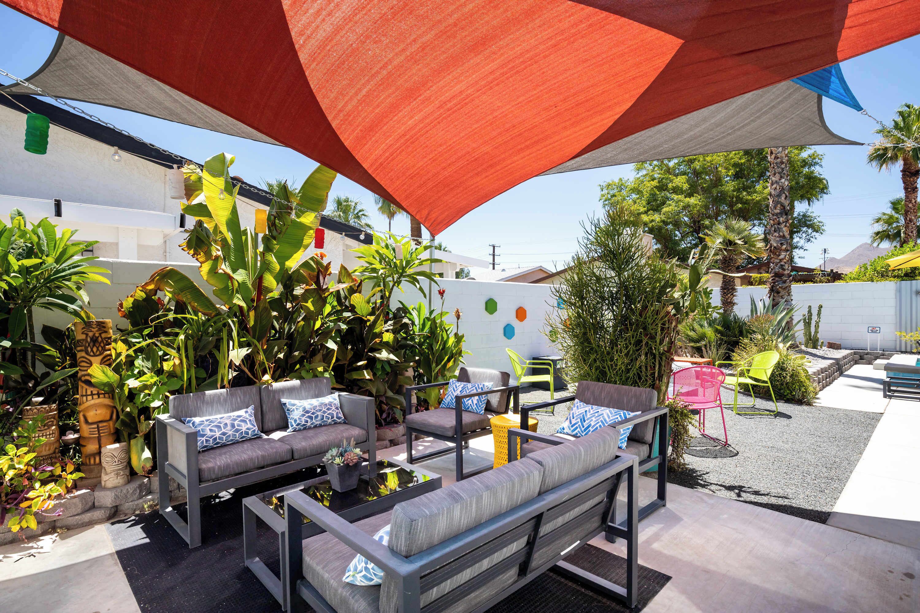 An outdoor seating area with grey sofas and chairs, surrounded by tropical plants and a large shade structure.