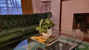 Una sala de estar con un sofá verde, una mesa de centro de vidrio con una planta y libros, y una chimenea.