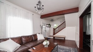 A spacious living room with a brown leather sofa, a wooden coffee table, and a chandelier. A staircase leads to a second floor, and a bench is visible in the background.