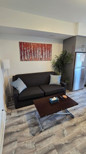 A living room with a dark sofa, a coffee table, a painting, a refrigerator, and a potted plant.