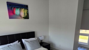 A bedroom with a bed, pillows, a nightstand, a lamp, and a wall art piece. - Minutes To The Falls:  Stylish Townhome Gem! (Niagara Falls)