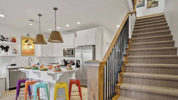 A modern kitchen with white cabinets, a central island, colorful stools, and a staircase with a wooden railing. - Live in Fantasy at Disney in the Desert! (Washington)