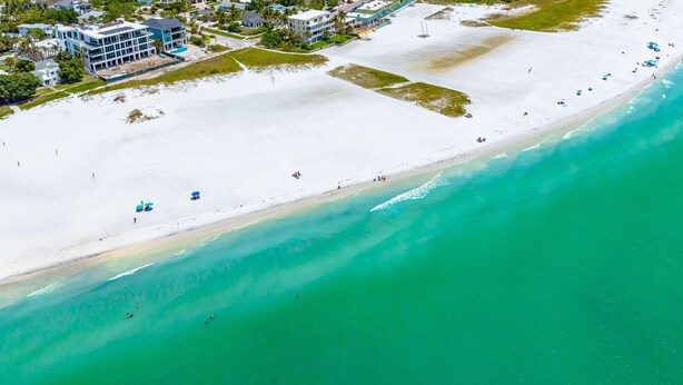 An aerial view of a beach with clear turquoise waters, white sand, and a few buildings in the background.