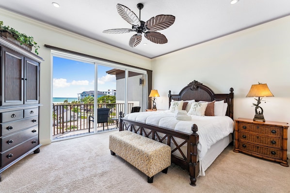 A bedroom with a large bed, a wooden dresser, a bench, a ceiling fan, and a view of the outdoors.
