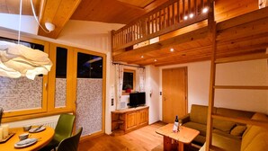 Ein gemütliches Zimmer mit Holzbalkendecke, einem Essbereich mit Tisch und Stühlen, einer Couch und einem Fernseher.
