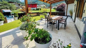 Ein überdachter Patio-Bereich mit Tisch und Stühlen, umgeben von Topfpflanzen und Blick auf eine gebirgige Landschaft.