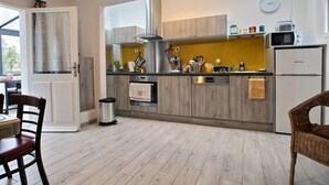 A modern kitchen with wooden cabinets, a stainless steel range hood, and a built-in oven. There is a dining area with a table and chairs, and a trash can is visible. - Le Gîte de Gravelodic (PAIMPOL)